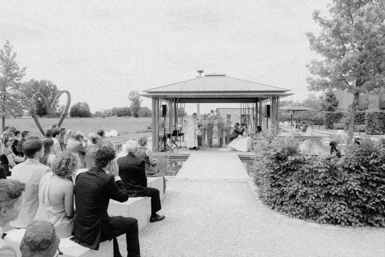 Sitzende Hochzeitsgäste mit Blick auf einen Holzpavillon am Wasser, in Schwarz-Weiß.