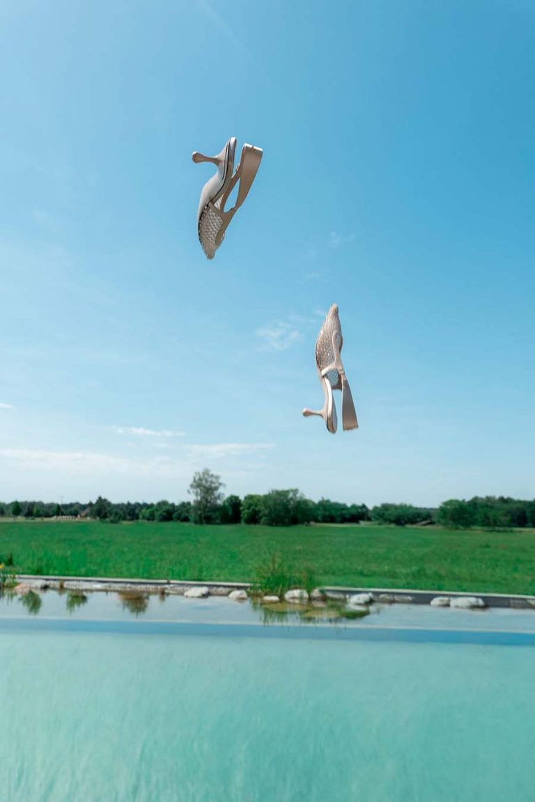 Zwei hochgeworfene Schuhe vor blauem Himmel über einer Wasserfläche und grünen Wiesen.