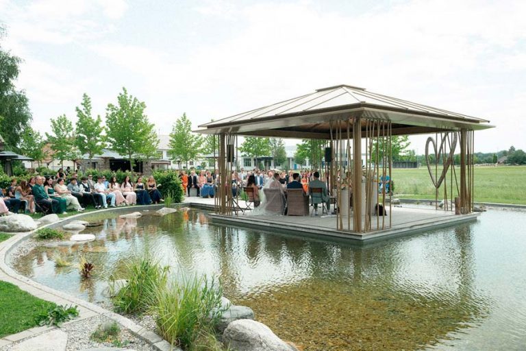 Holzpavillon mit Hochzeitsgesellschaft am Wasser und Spiegelung im See.