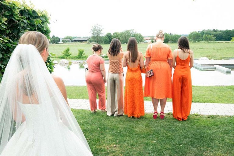 Braut mit langem Schleier vor einer Reihe von Bridesmaids in orangefarbenen Kleidern am See.