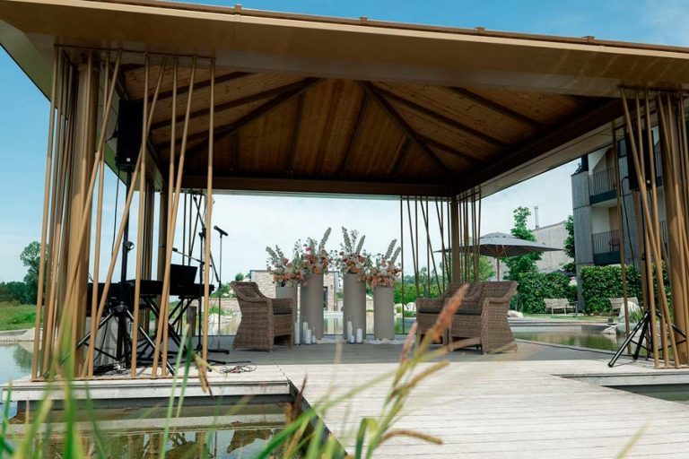 Offener Holzpavillon am Wasser mit heller Stoffdekoration und Blumenschmuck.
