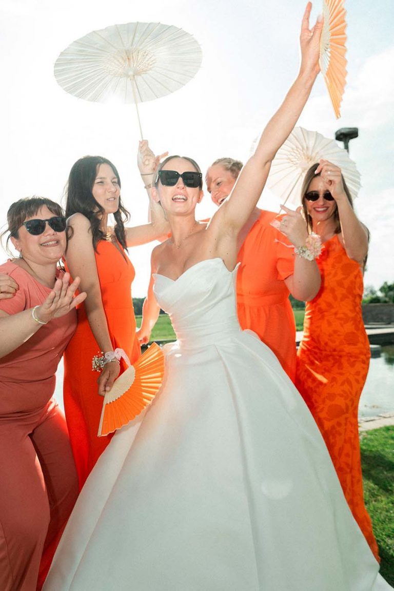 Braut mit Sonnenbrille und erhobenen Armen neben Bridesmaids in orangefarbenen Kleidern.