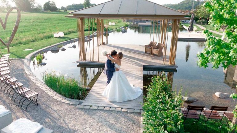 Brautpaar auf einem Weg am Wasser neben einem Holzpavillon, von oben fotografiert.
