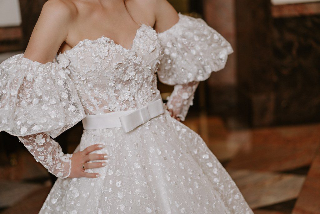 Detailaufnahme eines bestickten Brautkleids mit Puffärmeln und schmalem Gürtel in der Hotelhalle