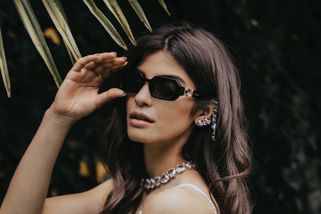 Porträt der Braut mit Sonnenbrille, Ohrschmuck und Kristallcollier vor dunklem Grün