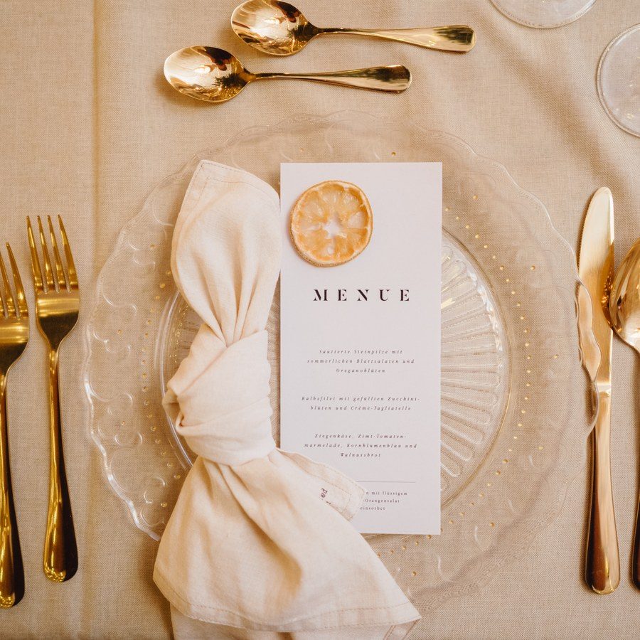 Eventplanung Bayern: Gedeck von oben mit goldenem Besteck, Organza-Serviette, Menükarte und Zitronenscheibe