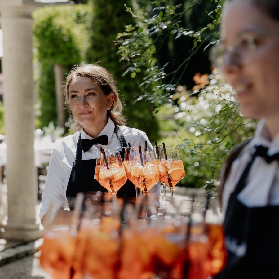 Eventplanung Ingolstadt: Service trägt Tabletts mit orangefarbenen Aperitif-Gläsern durch den Gartenempfang
