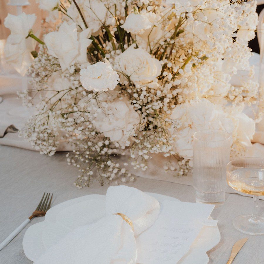 Eventplanung Mallorca: Gedeck mit Leinenserviette und goldenem Besteck vor einem Arrangement aus weißen Blüten