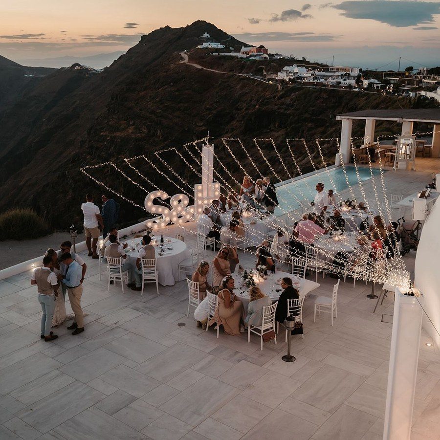 Eventplanung Santorini: Gäste feiern abends an runden Tafeln unter Lichterketten auf der Terrasse über dem Meer