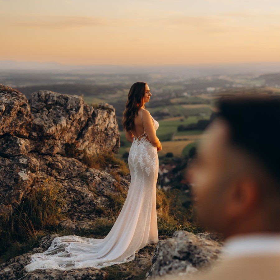 Freie Trauung Allgäu: Braut im Spitzenkleid steht auf einem Felsen und blickt im Abendlicht über die Hügellandschaft