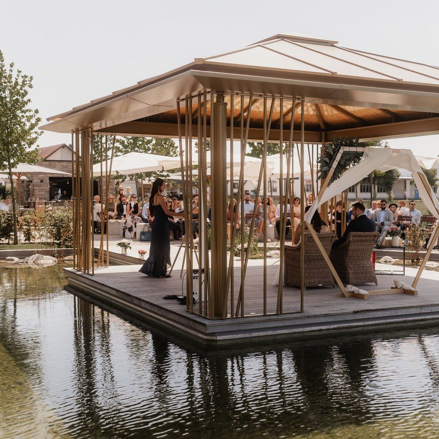 Freie Trauung bei einer Scheunenhochzeit: Brautpaar und Traurednerin auf einem Holzpavillon über dem Teich