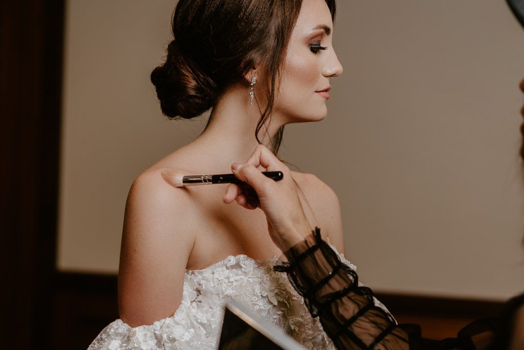 Braut wird am Hochzeitsmorgen geschminkt, während sie im bestickten Kleid auf dem Sessel sitzt