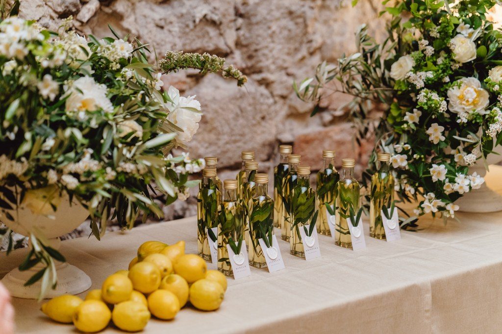 Kleine Flaschen als Gastgeschenke zwischen frischen Zitronen, Oliven­zweigen und weißen Blumen auf der Empfangstafel