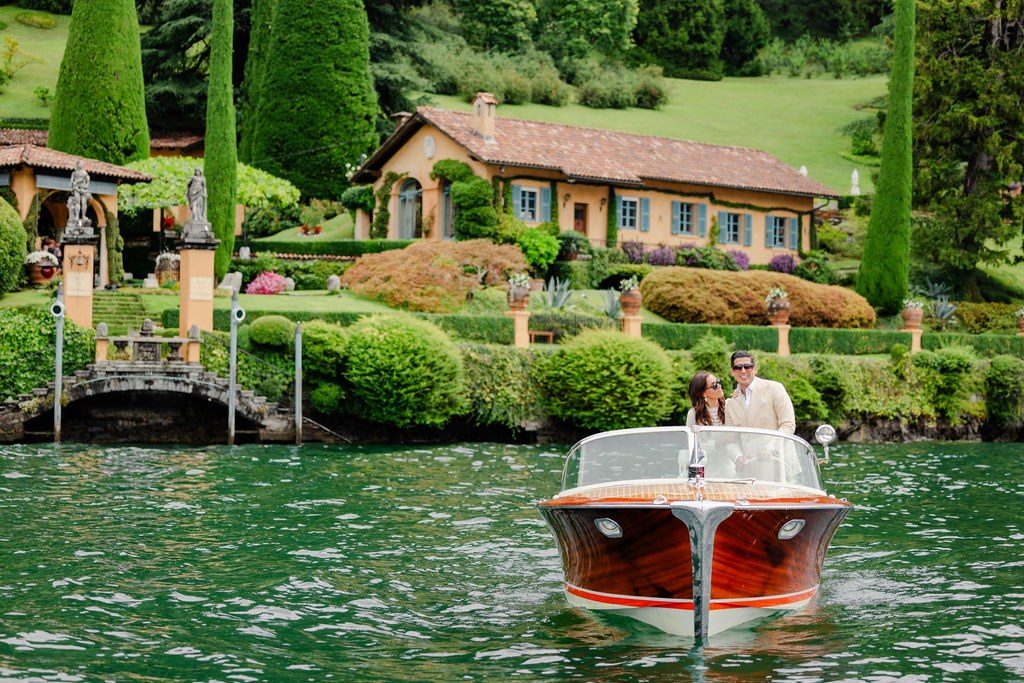 Brautpaar fährt in einem klassischen Holzboot über den See, dahinter eine Villa mit Zypressengarten
