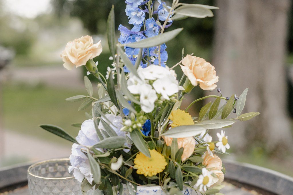Blumenarrangement in Blau, Gelb und Weiß in einer gemusterten Vase auf dem Holztisch, daneben Kumquats