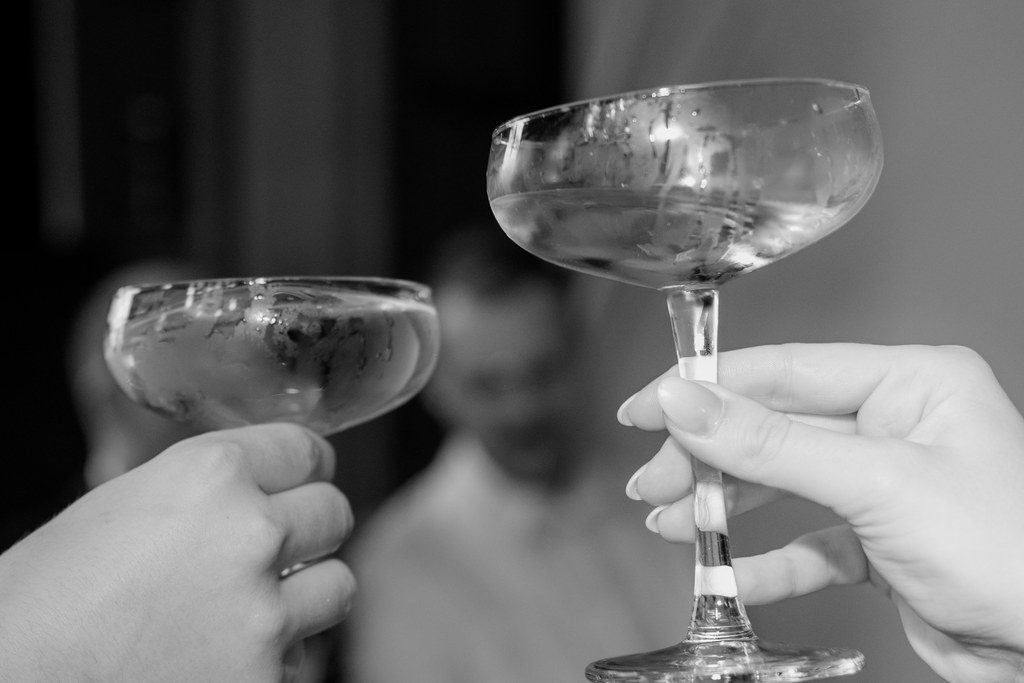 Zwei Hände stoßen mit Champagnerschalen an, im Hintergrund die feiernde Hochzeitsgesellschaft, Schwarz-Weiß