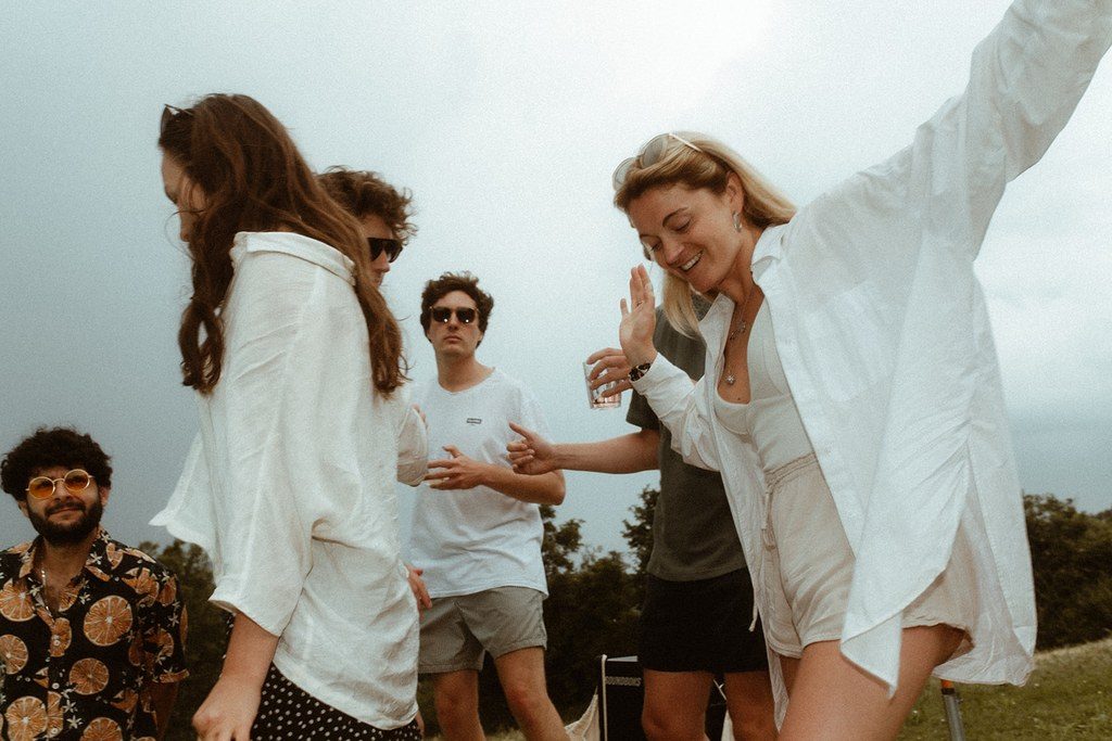 Hochzeitsgäste in Weiß tanzen und lachen im Sommerlicht auf der Wiese vor den Hügeln