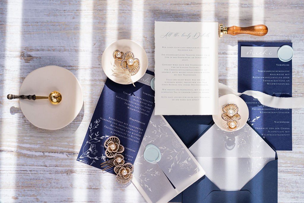 Hochzeitspapeterie in Dunkelblau und Gold mit Siegelstempel, Ringschale und Ohrringen auf hellem Holz