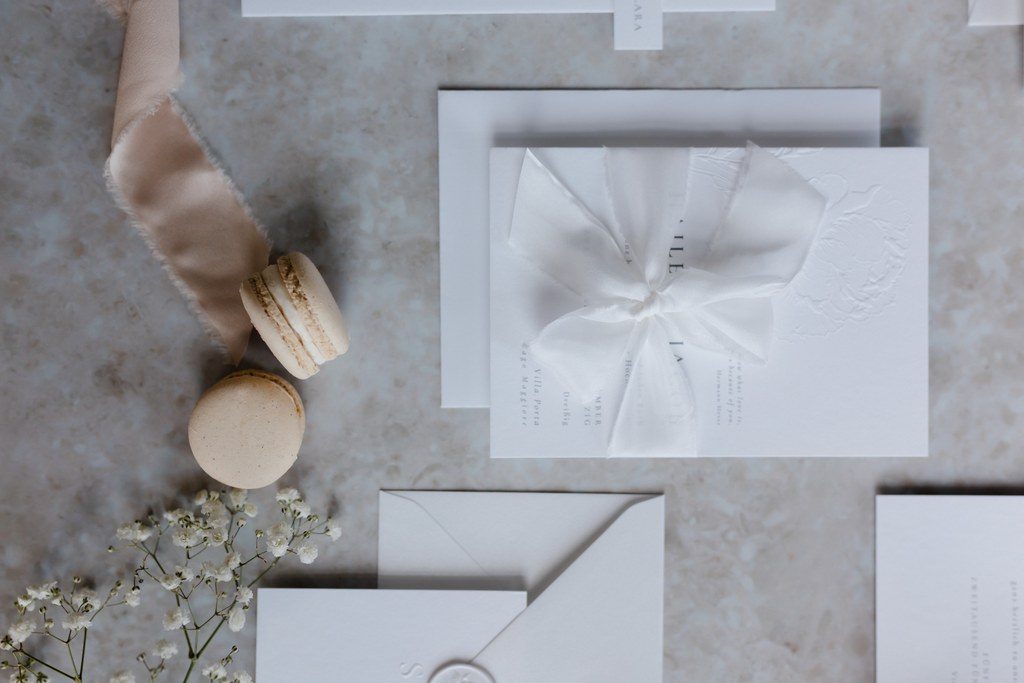 Weiße Hochzeitspapeterie mit Seidenband, Schleierkraut und Macarons auf hellem Marmor