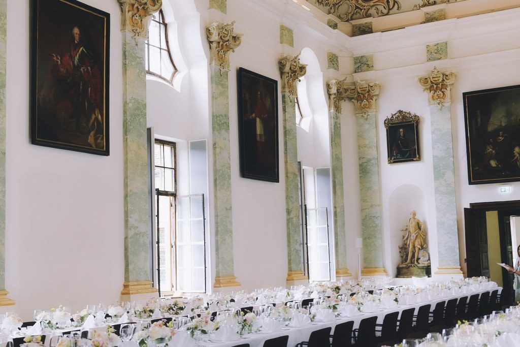 Lange Hochzeitstafel in einem barocken Saal mit Säulen, Gemälden und weißen Blumen