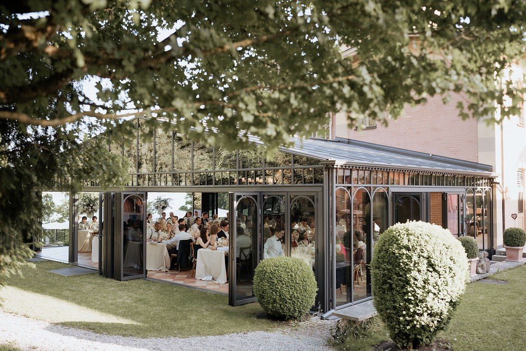 Gläserner Wintergarten einer Villa, in dem die Hochzeitsgäste an festlich gedeckten Tischen sitzen