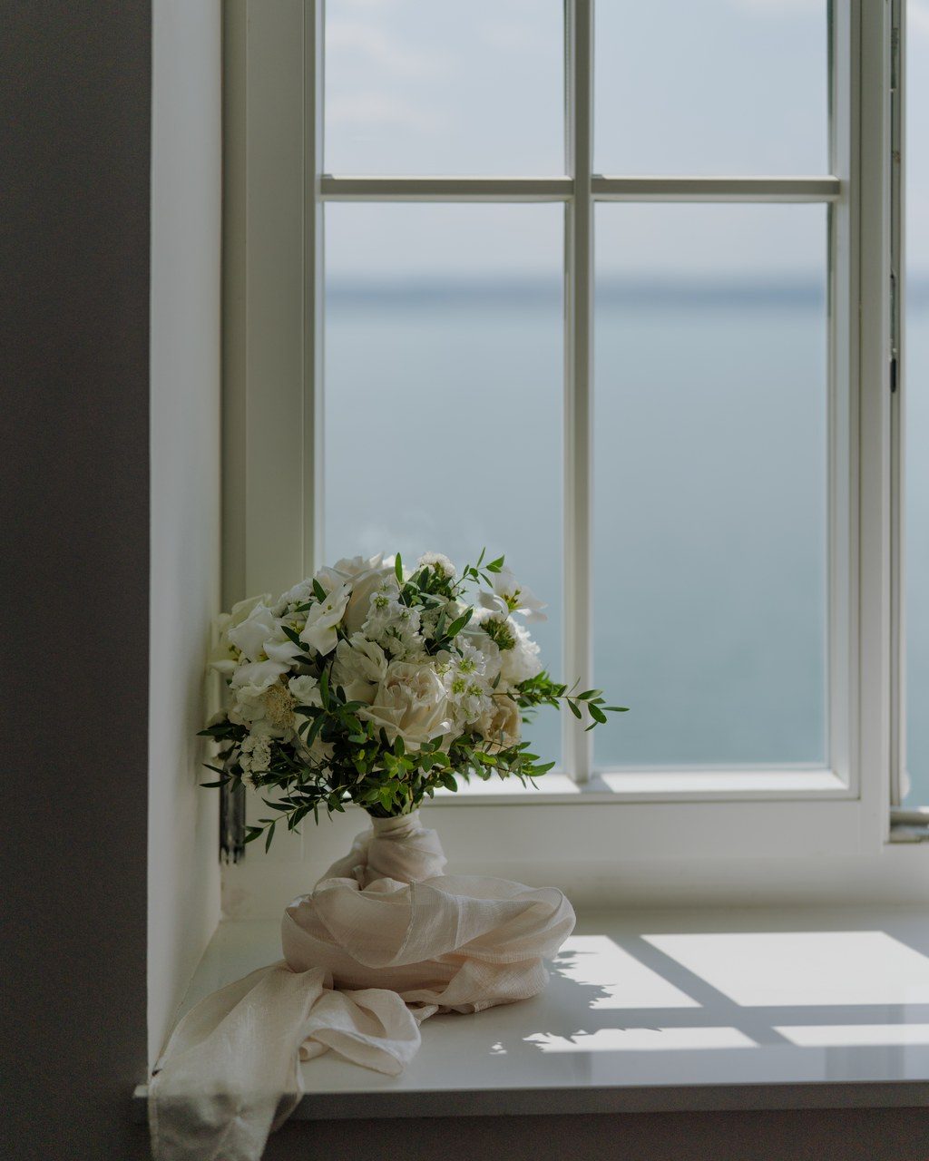 Hochzeitsplanerin Bodensee: weißer Brautstrauß liegt auf der Fensterbank, dahinter der stille See im Morgenlicht