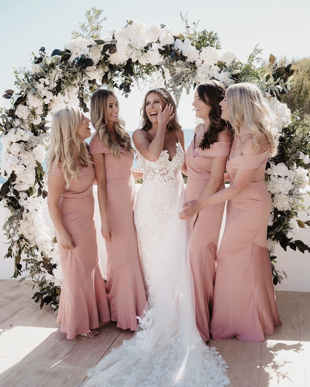 Hochzeitsplanerin Ibiza: Braut lacht mit ihren Brautjungfern in altrosa Kleidern unter dem Blumenbogen