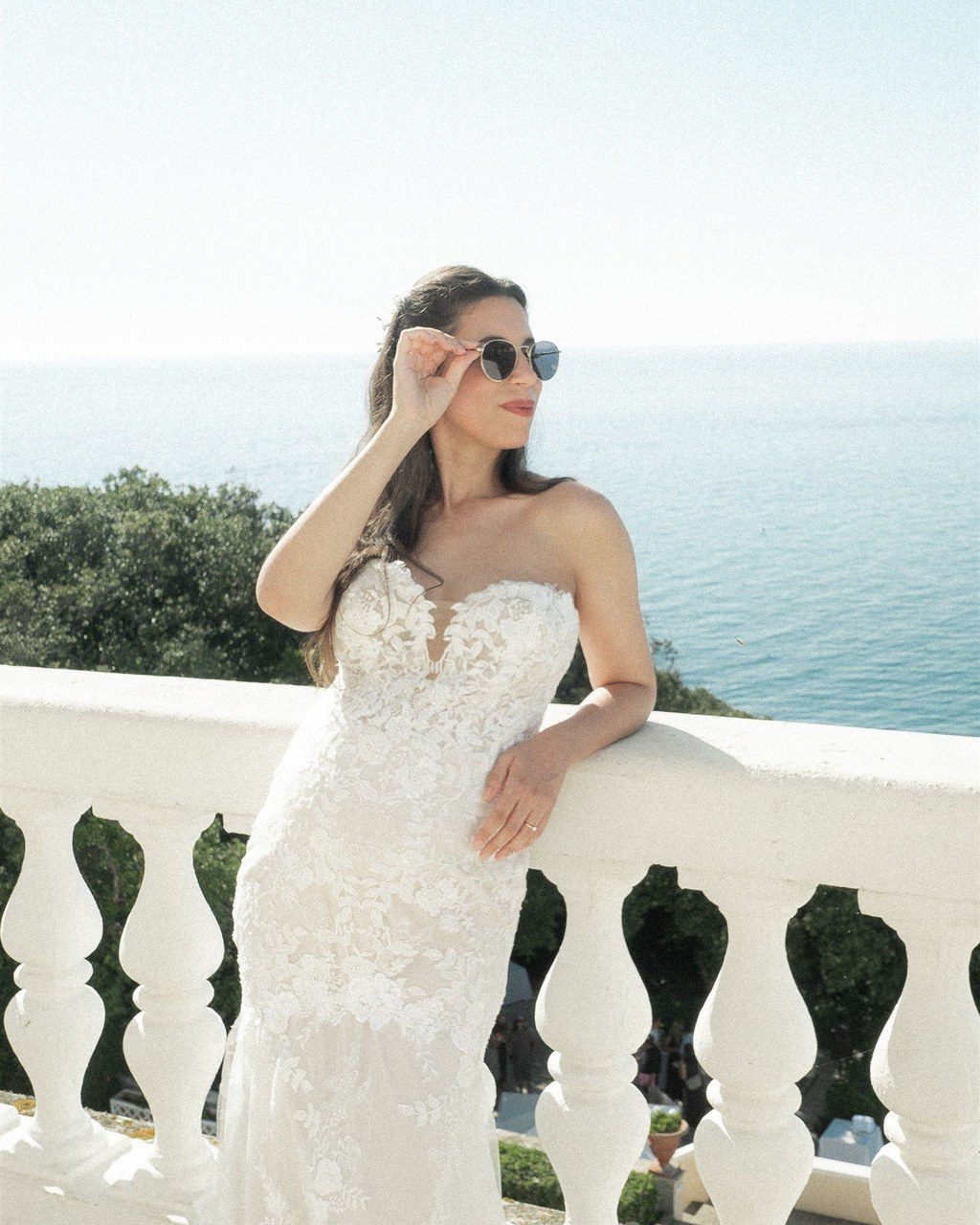 Hochzeitsplanerin Italien: Braut im Spitzenkleid lehnt mit Sonnenbrille an der Balustrade über dem Meer