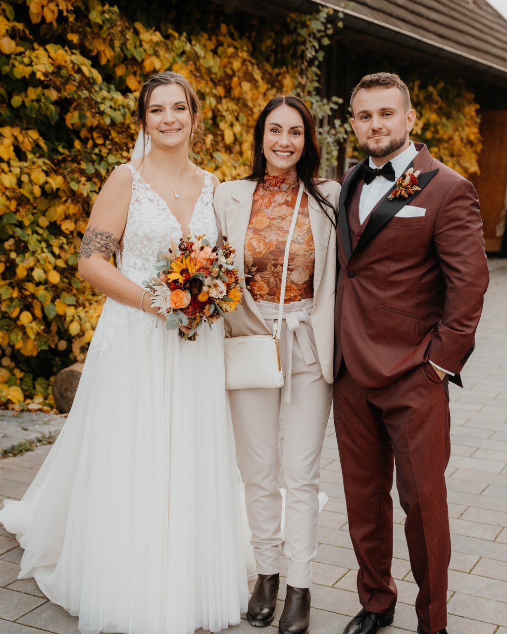 Hochzeitsplanerin Mannheim: Brautpaar mit herbstlichem Brautstrauß und Gast vor einer Wand aus gelbem Laub