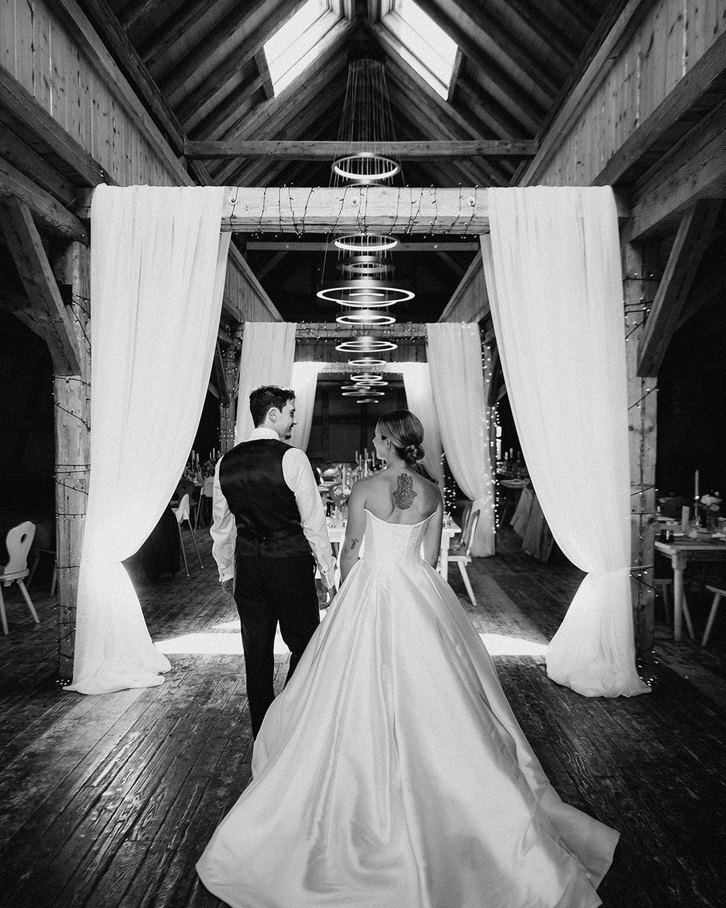 Hochzeitsplanerin Österreich: Brautpaar zieht durch weiße Vorhänge in die festlich beleuchtete Scheune ein, Schwarz-Weiß
