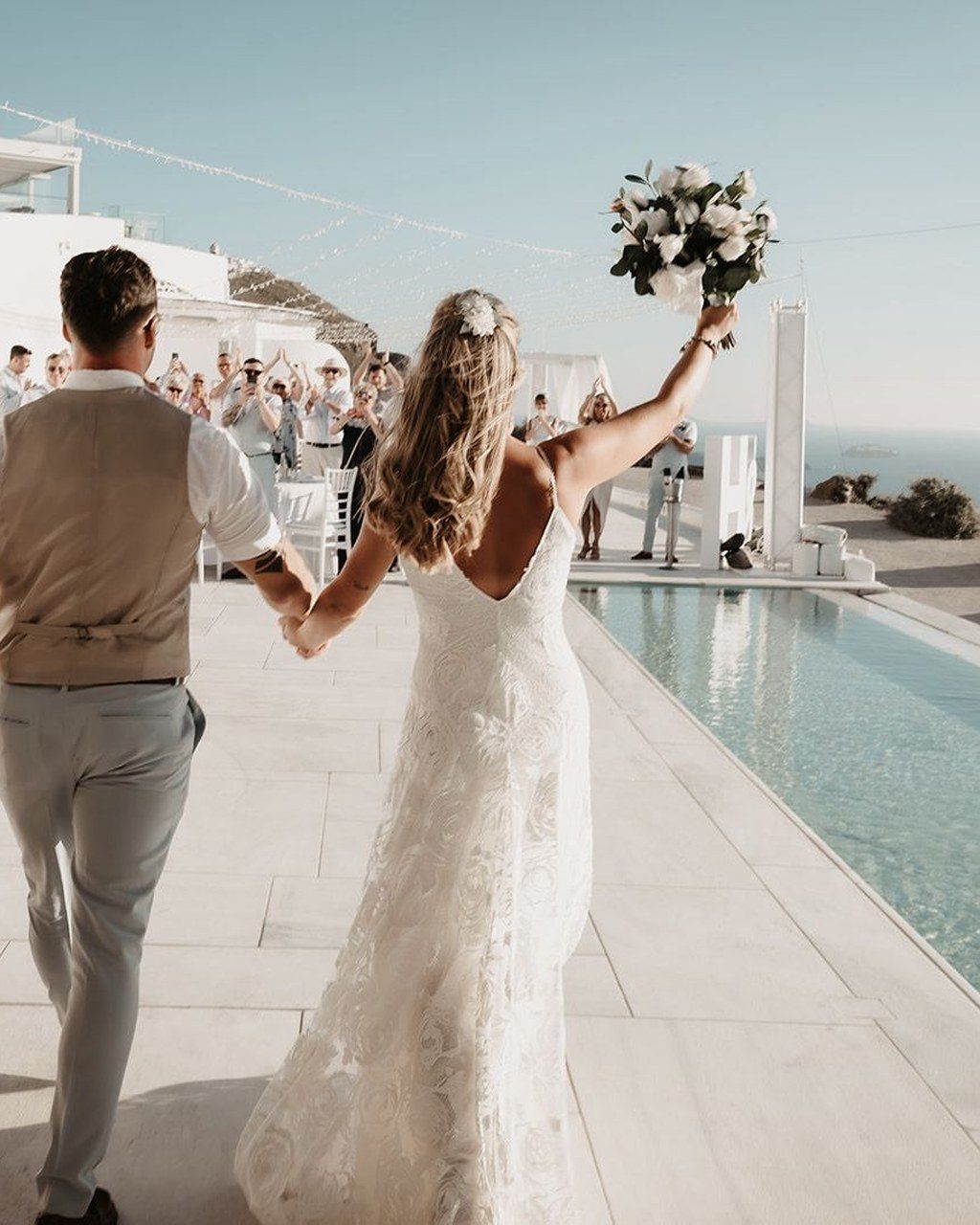 Hochzeitsplanerin Santorini: Braut reißt beim Einzug den Brautstrauß hoch, während die Gäste applaudieren