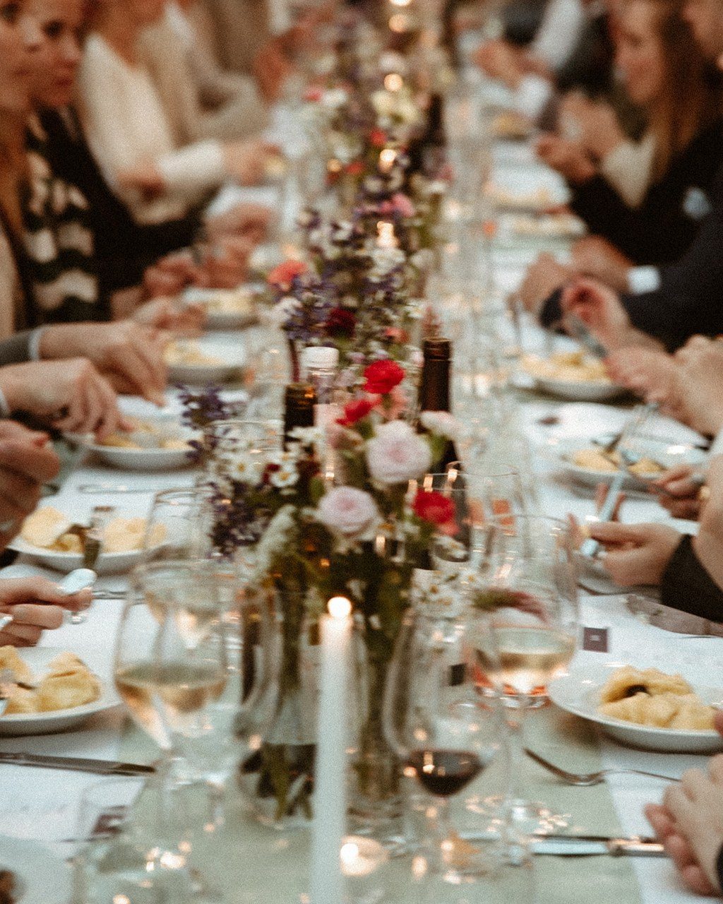 Hochzeitsplanerin Toskana: Gäste sitzen an der langen Tafel zwischen Kerzen, Weingläsern und kleinen Blumenvasen