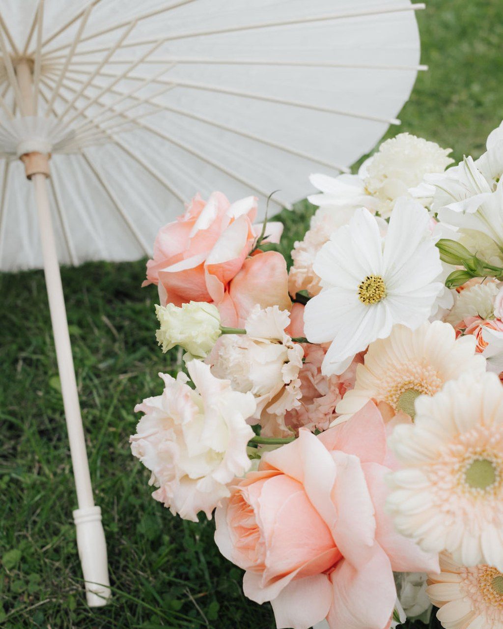 Weißer Papierschirm liegt im Gras neben Rosen, Cosmeen und Gerbera in Apricot und Weiß