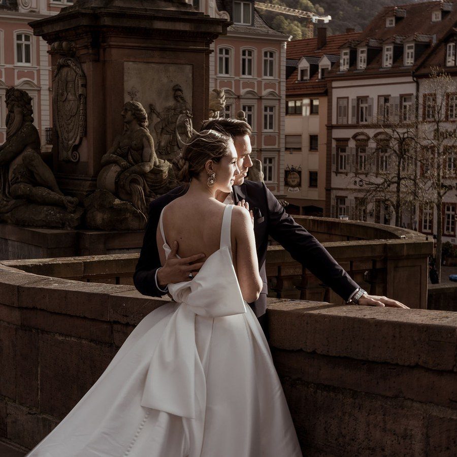 Hochzeitsplanung Heidelberg: Brautpaar steht an der Brüstung der Alten Brücke, im Hintergrund die Altstadtdächer und der Hang