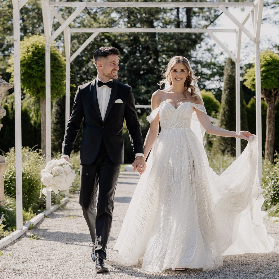 Hochzeitsplanung Ingolstadt: Brautpaar geht Hand in Hand durch eine weiße Pergola im Schlosspark