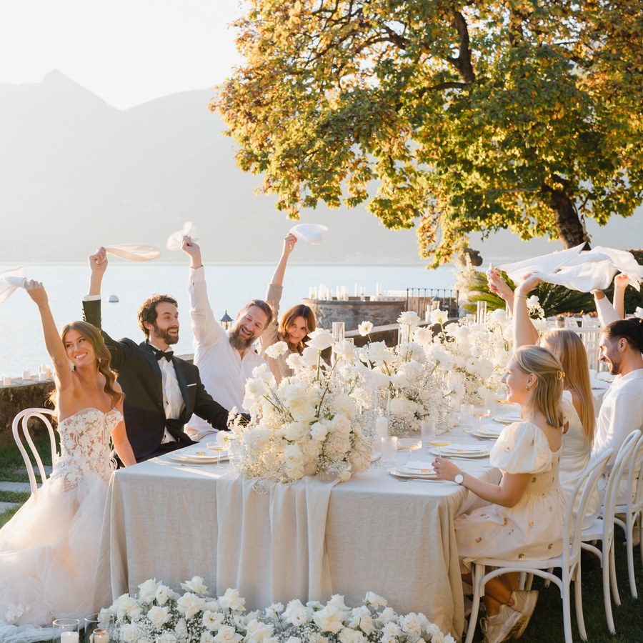 Hochzeitsplanung Lago Maggiore: Gäste jubeln an der weiß gedeckten Tafel im Garten direkt am Seeufer