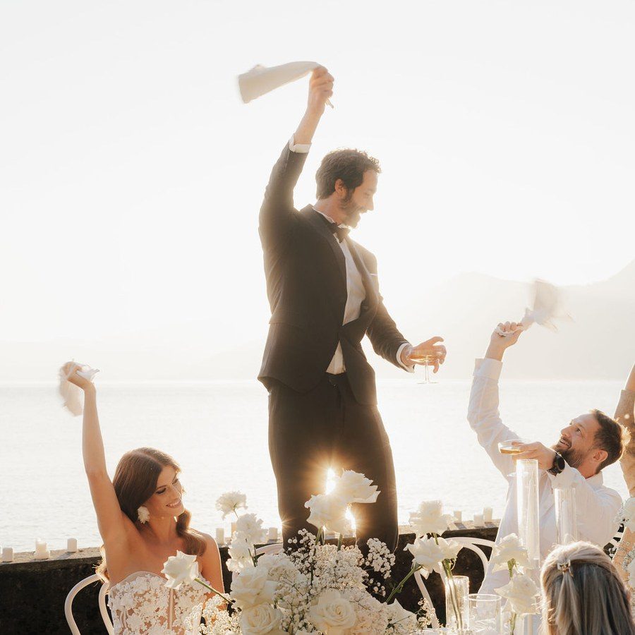 Hochzeitsplanung Mallorca: Gäste schwenken Servietten an der weiß gedeckten Tafel über dem Meer im Abendlicht