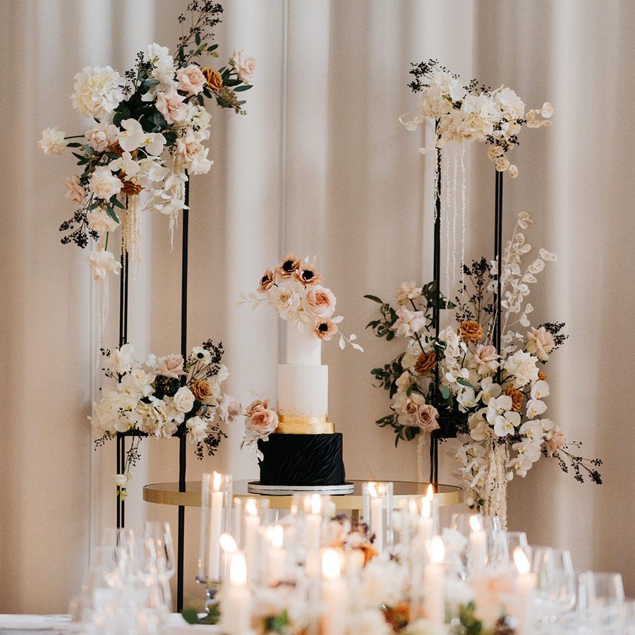 Hochzeitsplanung München: schwarz-weiße Hochzeitstorte auf goldenem Ständer zwischen Kerzen und hohen Blumenarrangements