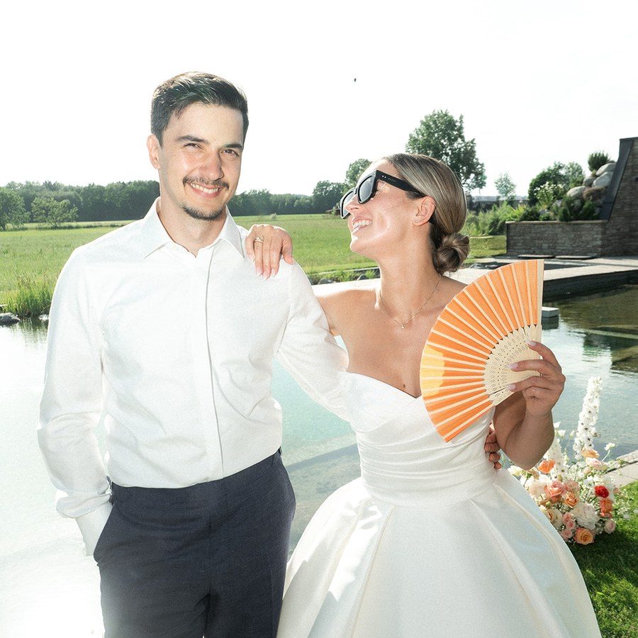 Hochzeitsplanung Österreich: Brautpaar steht lachend mit orangefarbenem Fächer am Naturteich