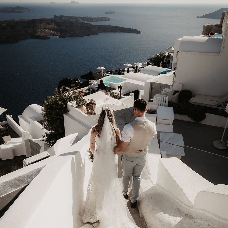 Hochzeitsplanung Santorini: Brautpaar geht mit langem Schleier über die weiße Terrasse hoch über der Caldera