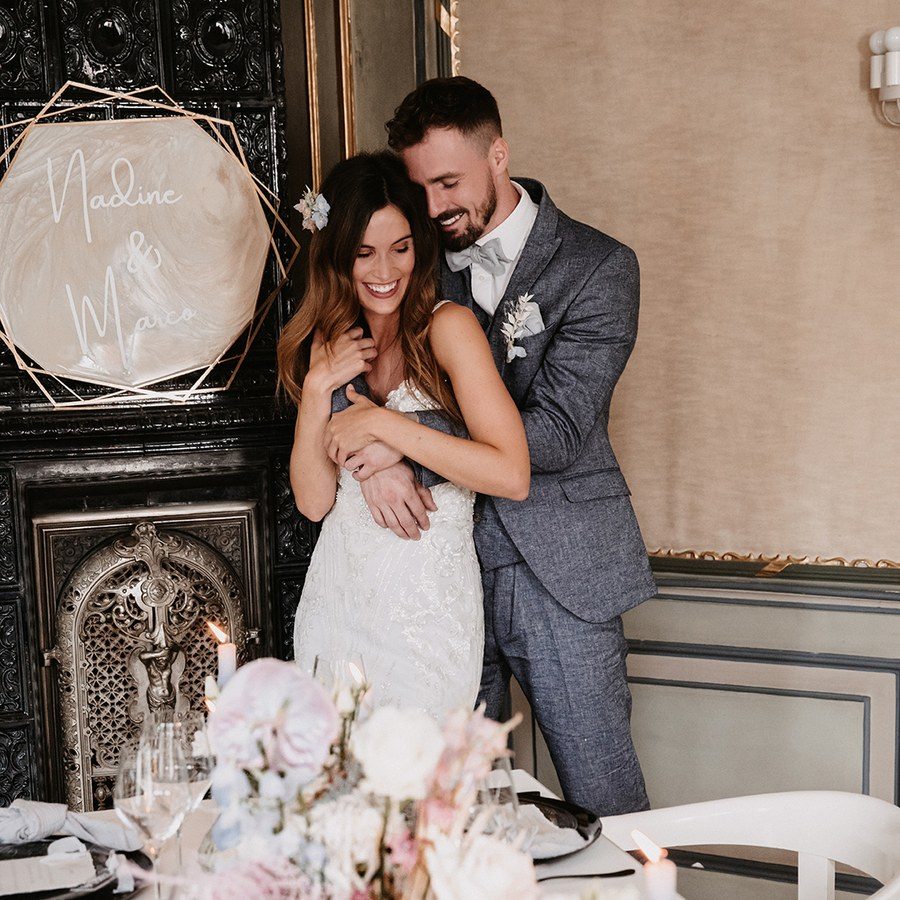 Hochzeitsplanung Stuttgart: Brautpaar umarmt sich im holzvertäfelten Saal, davor die Festtafel mit Blumen in Rosé und Hellblau