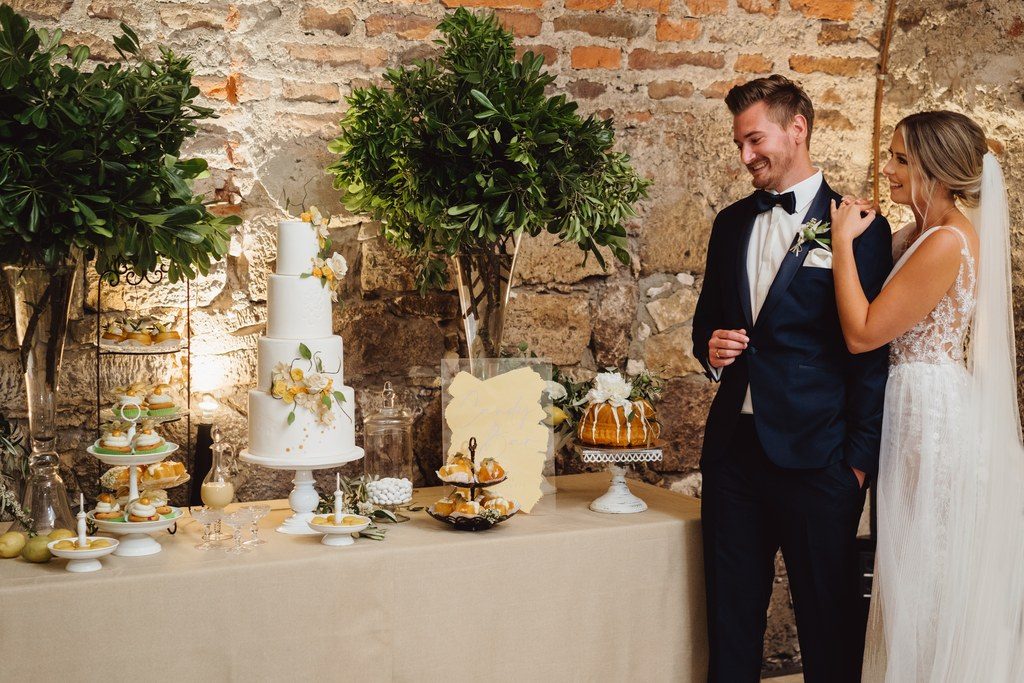 Brautpaar steht neben dem Sweet Table mit weißer Hochzeitstorte, Zitronentörtchen und Olivenbäumchen