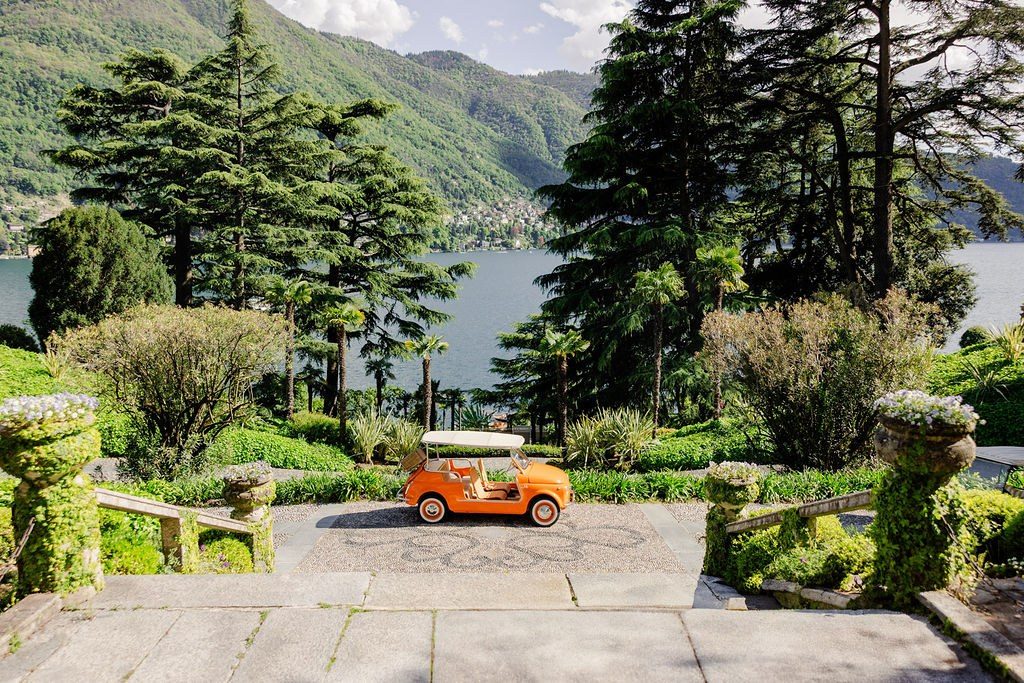Gartenterrasse mit Steinbalustraden, einem kleinen orangefarbenen Oldtimer und Blick auf See und Berge