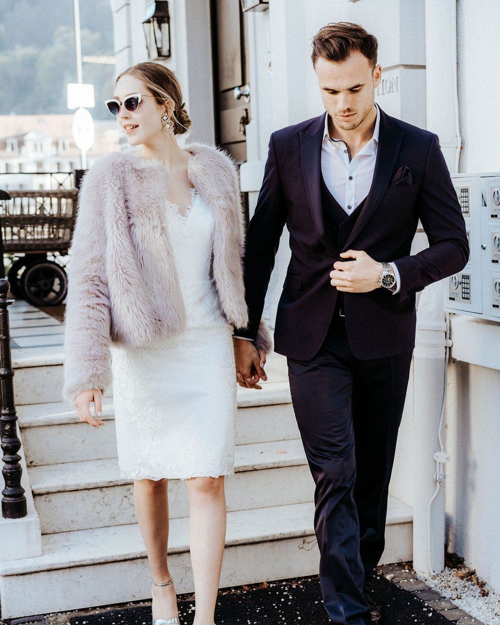 Braut im kurzen Kleid mit rosafarbenem Mantel und Sonnenbrille geht Hand in Hand mit dem Bräutigam eine Altstadttreppe hinunter