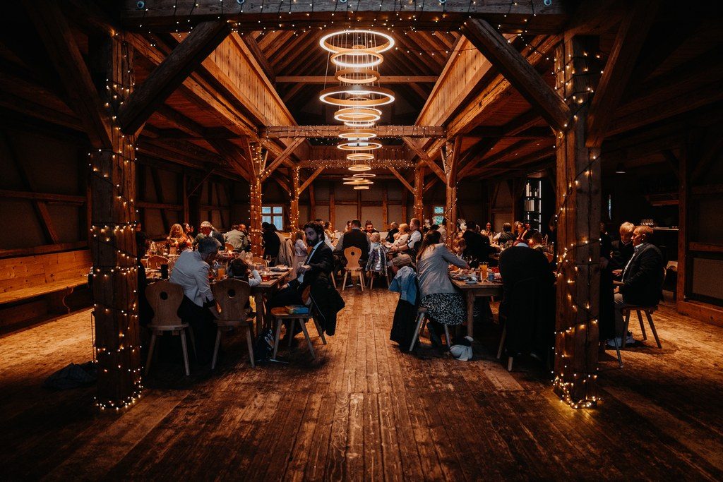 Hochzeitsdinner in einer Holzscheune mit Lichterketten und runden Leuchtringen unter dem Dach
