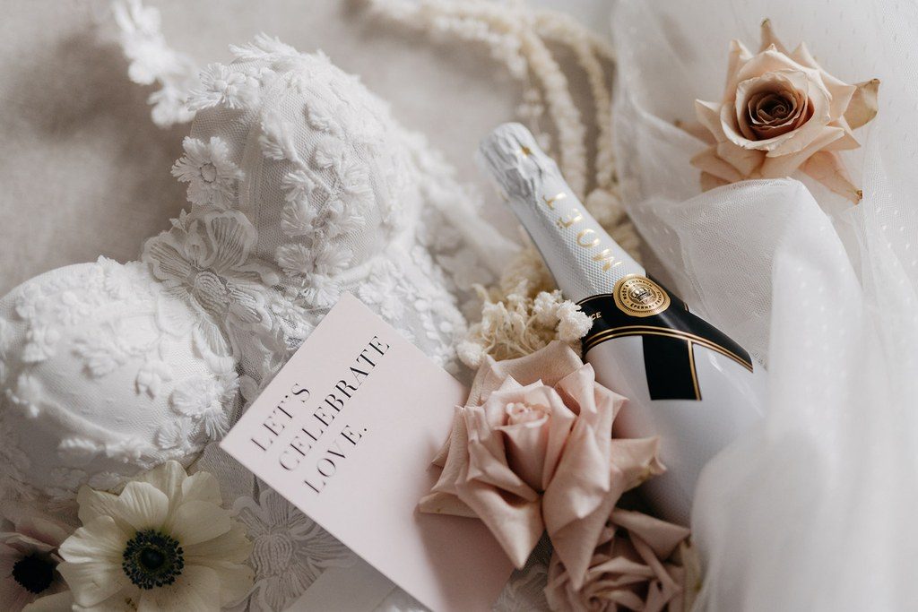 Champagnerflasche mit Rosen, Anemonen und Karte neben dem Spitzendetail des Brautkleids am Hochzeitsmorgen